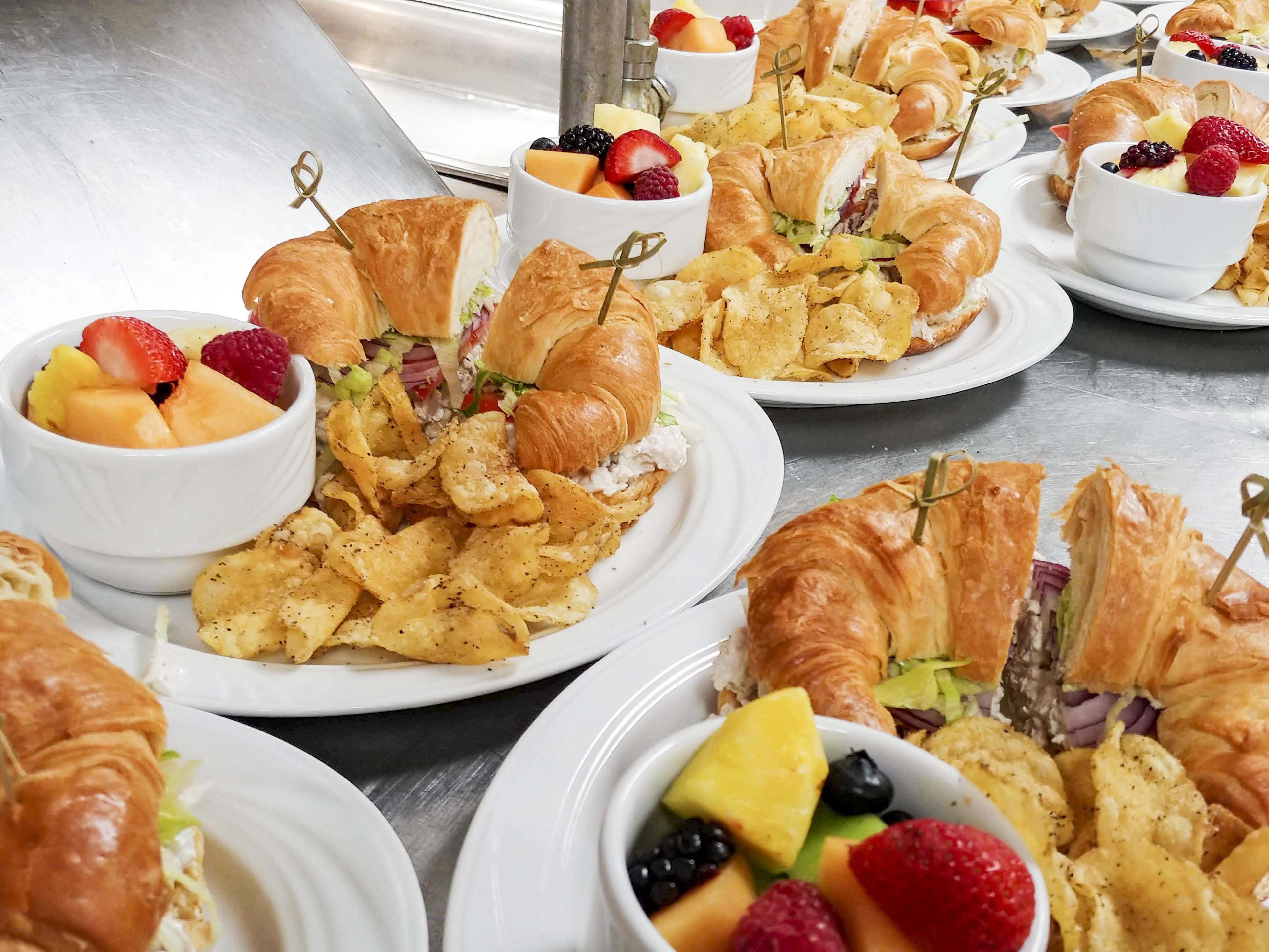 Close up photo of plates of sandwiches, fruit, and chips.